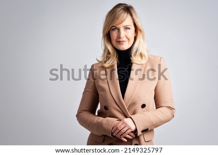 Studio portrait of attractive blond haired woman wearing turtleneck sweater and blazer while standing at isolated grey background. 