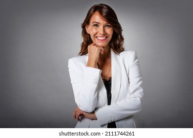 Studio portrait of attractive brown haired woman wearing white blazer and laughing while standing at isolated grey background. Copy space. Hand on chin.