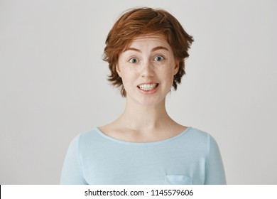 Studio Portrait Of Attractive Caucasian Female With Red Hair And Freckles Feeling Awkward, Lifting Eyebrows And Smiling Nervously, Having No Clue What To Say In Strange Situation On Meeting