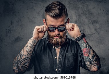 Studio portrait of a bearded hipster man with tattoos on his arms and neck  dressed in a black shirt and sunglasses