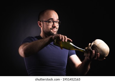 Studio Portrait Of A Bearded Man In A Blue T-shirt And Glasses, Photographed Against A Black Background. A Man With A Sly Look Pours Alcohol From A Bottle Into The Mouth Of The Skull