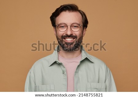 studio portrait of bearded man posing over beige background looking into camera with broad smile on his face