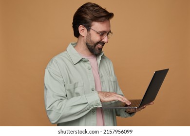 Studio Portrait Of Bearded Man Standing Over Beige Background Wears Casual Shirt And Glasses Typing Message On Laptop Computer, Smiles Broadly