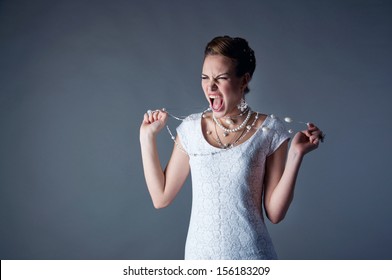Studio portrait of beautiful angry and agressive shouting bride wearing white dress, screaming and crying at someone, trying to tear her pearl beads apart against gray background. Copy space