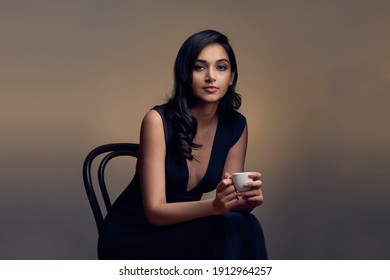 Studio Portrait of a beautiful, elegantly dressed Indian Asian woman in a black evening dress holding a small white cup of coffee. She is sitting in a photography studio and is gazing into the camera.