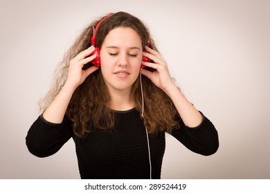 Studio portrait of a beautiful ginger teenage girl with long curly hair listening to music on red headphones isolated against a grey background