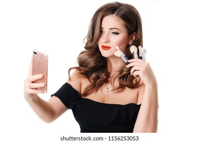 Studio Portrait Of Beautiful Trendy Girl With Long Curly Hair And Make Up Holding Arm On Waist And Taking Selfie Via Cell Phone Over White Isolated Background. 