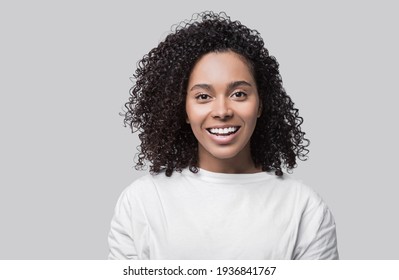 Studio Portrait Of A Beautiful Young Woman With Black Curly Hair. Laughing Mixed Race Girl Wearing White T-shirt Looking At Camera. Isolated On Grey Background. People, Lifestyle, Beauty Concept