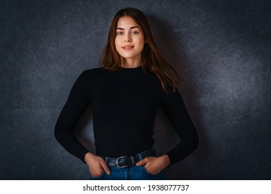 Studio Portrait Of Beautiful Young Woman Looking At Camera And Smiling While Standiing At Isolated Dark Background. 