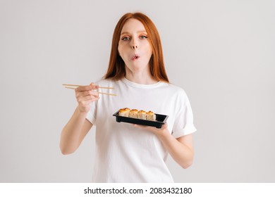 Studio Portrait Of Beautiful Young Woman Eating Delicious Sushi Rolls With Chopsticks Looking At Camera Standing On White Isolated Background. Pretty Caucasian Redhead Female Chewing Asian Food.