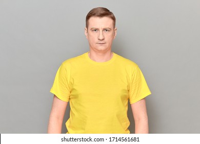 Studio portrait of blond mature man wearing yellow T-shirt, with neutral expression on face, with calm facial emotion, holding hands along body, standing over gray background