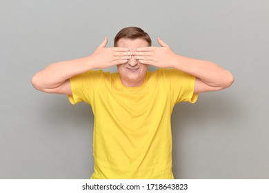 Studio Portrait Of Blond Mature Man Wearing Yellow T-shirt, Covering Eyes With Hands, Not Wanting To See Something Or Waiting Surprise, Standing Over Gray Background