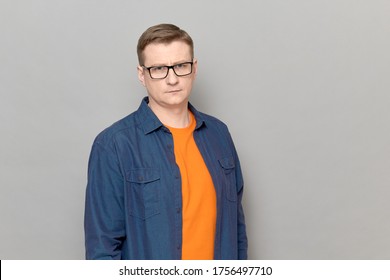 Studio portrait of blond mature man with glasses, wearing casual blue shirt, with serious expression on face, looking confident, holding arms along the body, standing over gray background