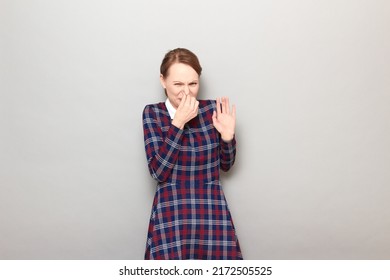 Studio portrait of blond young woman pinching her nose with hand, wincing in disgust, not wanting to sniff stinky nasty smell, wearing checkered dress, standing over gray background