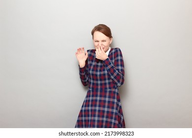 Studio portrait of blond young woman pinching her nose with hand, wincing in disgust, not wanting to sniff stinky nasty smell, wearing checkered dress, standing over gray background