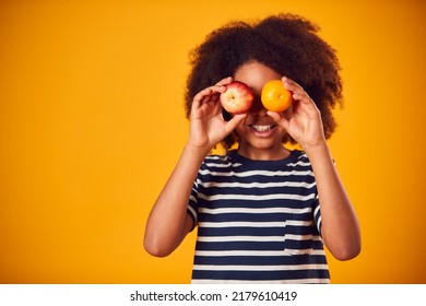 Studio Portrait Of Boy Holding Apple And Orange In Front Of Eyes Against Yellow Background