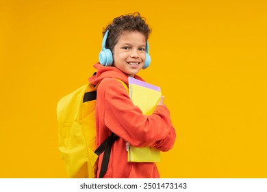 Studio portrait of cheerful African American schoolboy wearing orange hoodie, bright backpack and wireless headphones, posing over yellow background with a books pile in hands. - Powered by Shutterstock