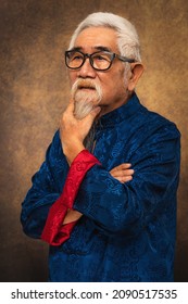 Studio Portrait Of Chinese Old Man Wearing Eyeglasses With White Hair And Beard In Tradional Chinese Clothing On Studio Backdrop Background