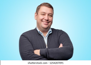 Studio portrait of confident plump european mature man smiling and crossing hands on chest standing on blue background.