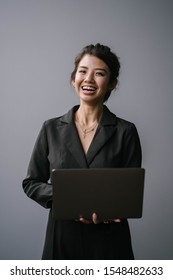 Studio Portrait Of A Confident, Young, Professional And Attractive Business Woman In A Black Dress Smiling As She Looks Up From Typing On Her Laptop Computer Against A White / Silver Background. 