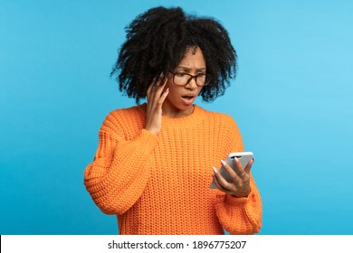 Studio Portrait Of Confused Shocked African Ethnicity Woman Looking At Smartphone With Opened Mouth, Reading Bad News, Notification, Unpleasant Message Isolated On Blue Color Background.