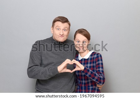 Studio portrait of couple making heart gesture with fingers, looking funny and crazy, making goofy faces, standing together over gray background. Relationship concept, human emotions and love