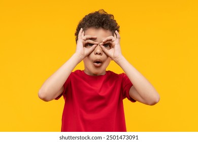 Studio portrait of cute little African American boy having fun making superhero's mask or imaginary binocular with his fingers, isolated over bright colored orange yellow background. - Powered by Shutterstock