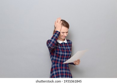 Studio portrait of disappointed young woman wearing checkered dress, holding paper, reading document, slapping her forehead with hand, realizing fatal mistake she made, standing over gray background