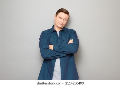 Studio portrait of disgruntled blond mature man holding arms crossed on chest, looking tired and bored, wearing casual blue shirt, standing over gray background - Shutterstock ID 2114230493