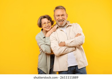 Studio portrait of elderly couple posing on yellow background, senior handsome man and pretty woman embracing, smiling at camera