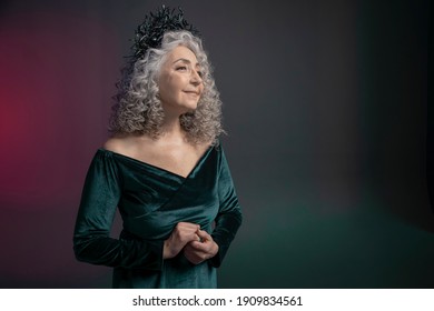 Studio Portrait Of An Elderly Woman 60-65 Years Old In A Beautiful Long Dress With A Crown Of Snakes On Her Head On A Colored Background. Concept: Stylish Pensioners Of Model Appearance, Active Life.