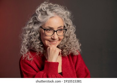 Studio portrait of an elderly woman 60-65 years old in a red sweater and glasses, gray curly long hair, on a colored background. Concept: stylish pensioners of model appearance, active life.