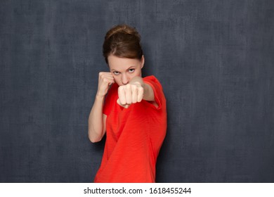 Studio portrait of enraged dissatisfied blond girl wearing orange T-shirt, holding fists clenched in front of her, making punch, ready to fight and defend her rights, standing over gray background