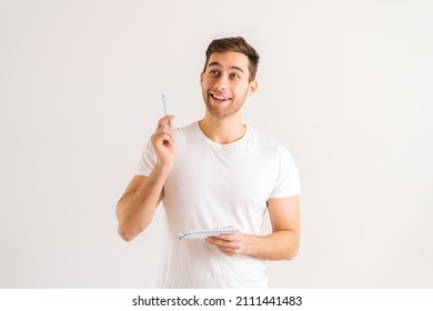 Studio Portrait Of Enthusiastic Young Man Writing In Copybook, Raising Pen Up Eureka Gesture On White Isolated Background. Happy Male Studying, Preparing For Exam, Making Notes In Paper Notebook.