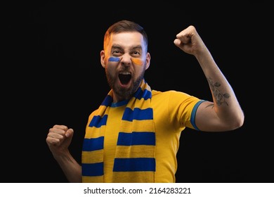 Studio Portrait Of Euphoric Happy Soccer Fan Man In Yellow-blue T-shirt And Scarf And Painted Face, Cheering For His Favourite Team. Isolated Over Black Background.