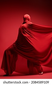 Studio Portrait Of An Expressive Female Fashion Model Wrapped In Red Fabric Posing On A Red Background. High Fashion Shoot. Full Length Portrait.