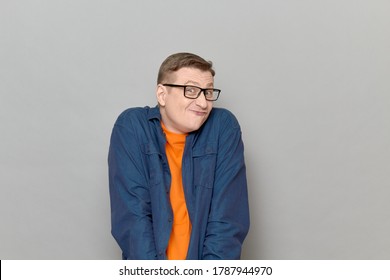 Studio Portrait Of Funny Blond Mature Man With Glasses, Wearing Casual Blue Shirt, Looking Like Stupid Bewildered User Or Shy Confused Nerd, Making Goofy Grimace, Standing Over Gray Background