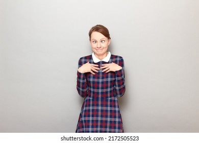 Studio Portrait Of Funny Blond Young Woman Making Silly Goofy Face, Grimacing With Crossed Eyes, Looking Confused And Perplexed, Wearing Checkered Dress, Standing Over Gray Background