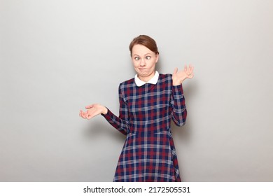 Studio Portrait Of Funny Blond Young Woman Making Silly Goofy Face, Grimacing With Crossed Eyes, Looking Confused And Perplexed, Wearing Checkered Dress, Standing Over Gray Background