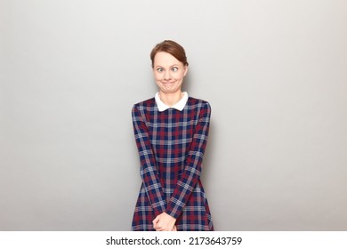 Studio Portrait Of Funny Blond Young Woman Making Silly Goofy Face, Grimacing With Crossed Eyes, Looking Confused And Perplexed, Wearing Checkered Dress, Standing Over Gray Background