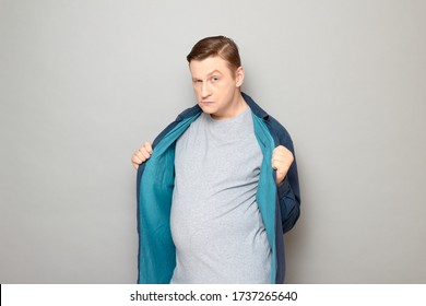 Studio Portrait Of Funny Comical Blond Mature Man Opening Blue Shirt And Showing His Tummy, Trying To Seem Attractive, Seductive And Sexy, Flirting, Standing And Posing Over Gray Background