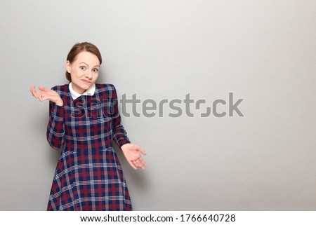 Studio portrait of funny confused blond girl wearing checkered dress, grimacing, waving hands, shrugging shoulders, not understanding how this happened, standing over gray background, with copy space