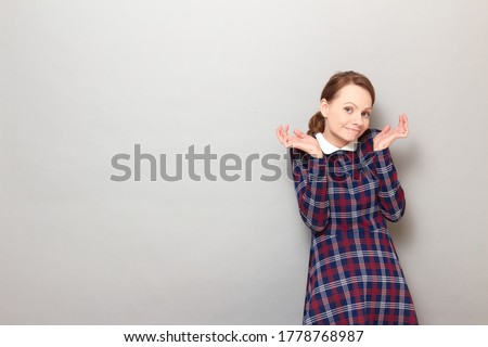 Studio portrait of funny confused perplexed blond girl wearing checkered dress, making silly goofy face, raising hands, shrugging shoulders, standing over gray background, with copy space