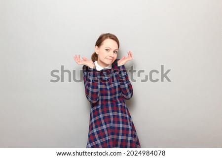 Studio portrait of funny confused perplexed blond young woman wearing checkered dress, making silly goofy face, raising hands, saying oops, shrugging shoulders, standing over gray background
