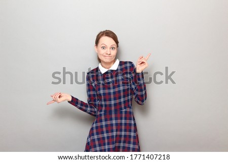 Studio portrait of funny confused young blond woman wearing checkered dress, looking bewildered and puzzled, doubting what to choose, feeling perplexity, standing over gray background