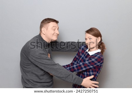 Studio portrait of funny couple, happy cheerful man is trying to hug woman resisting and grimacing from displeasure, both are standing over gray background. Relationship concept, human emotions