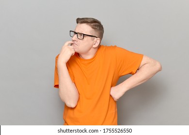 Studio portrait of funny focused puzzled blond mature man with glasses, wearing T-shirt, touching lips with finger, thinking hard, trying to understand something, standing over gray background