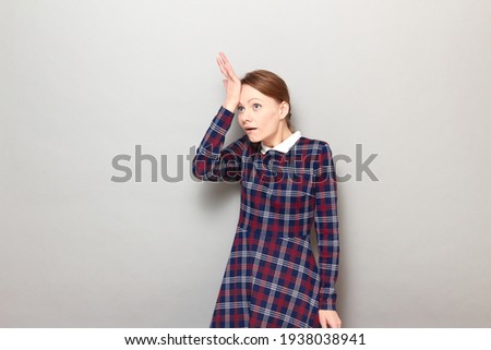 Studio portrait of funny goofy young blond woman wearing checkered dress, slapping her forehead with hand, realizing silly mistake she made, saying duh, standing over gray background