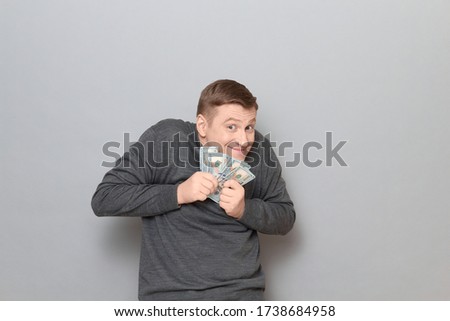Studio portrait of funny happy mature man wearing casual jumper, holding bunch of US dollars in hands, clasping money to his chest, looking greedy and sly, standing hunched over gray background