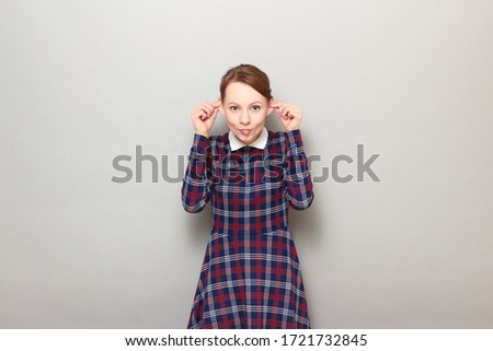 Studio portrait of funny happy young blond woman wearing checkered dress, touching ears with hands and sticking out her tongue, smiling, fooling around and having fun, standing over gray background